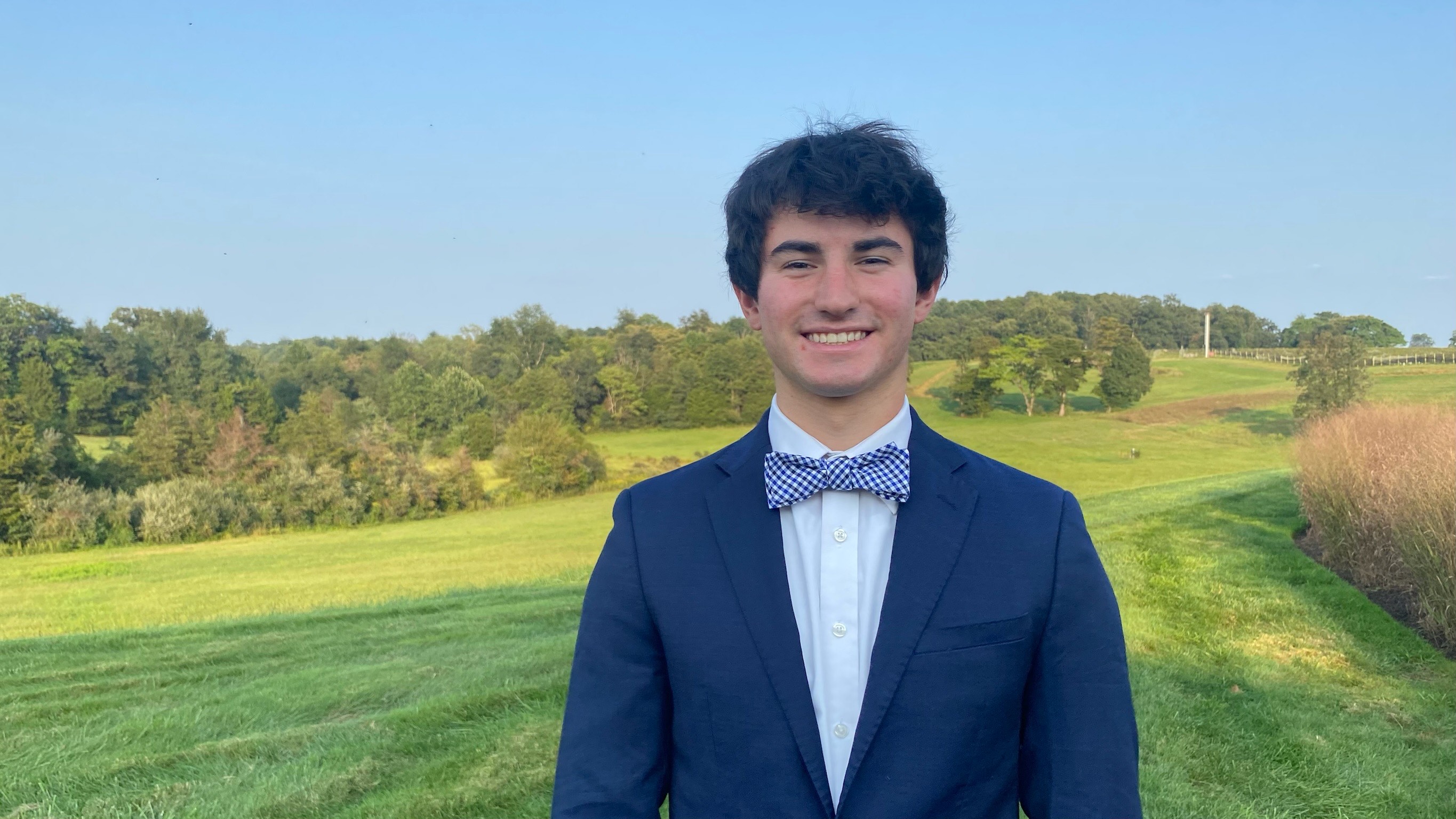 A smiling young man stands on a green golf course in a blue suit and bowtie.