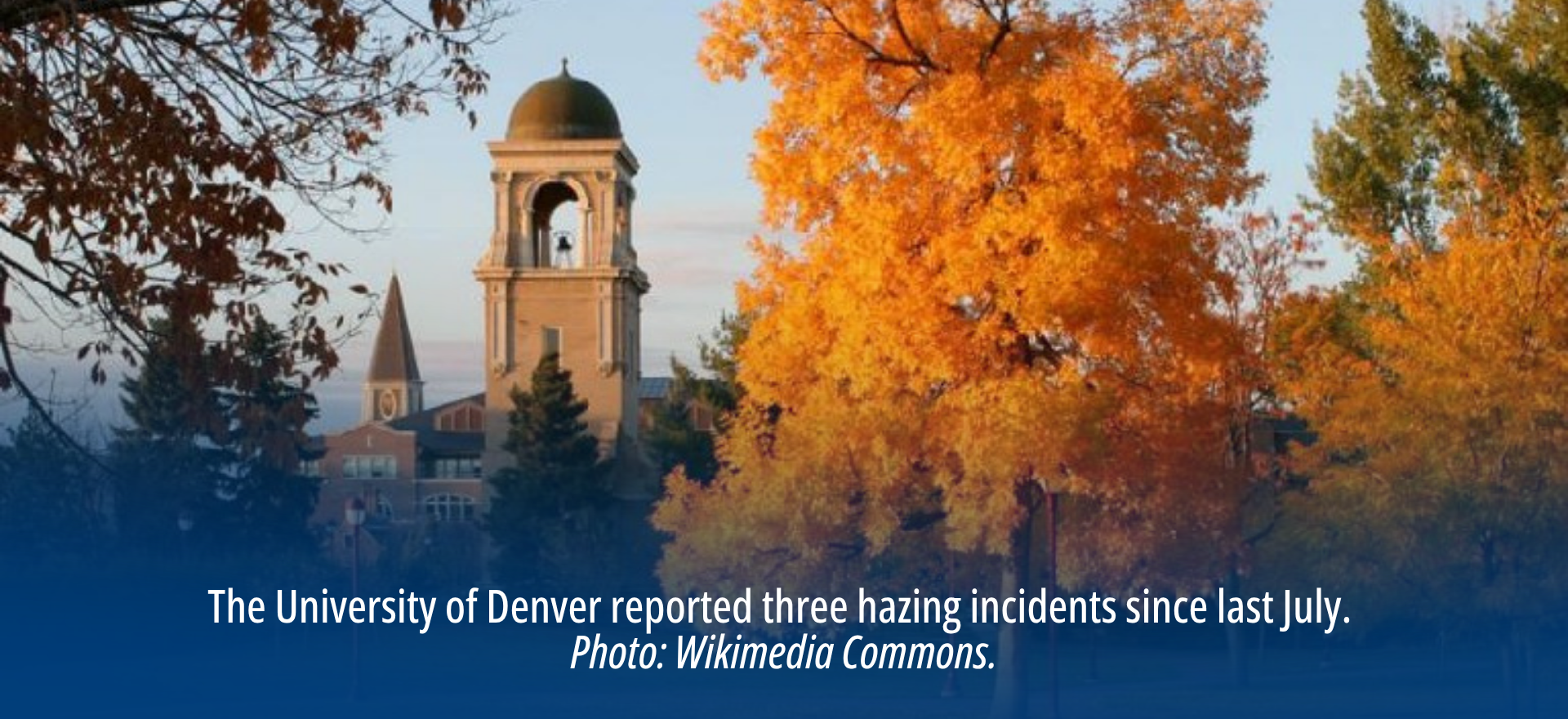 A bell tower on a college campus stands near large trees, some of which have orange leaves.